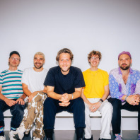 Fünf Musiker der Band Il Civetto aus Berlin sitzen auf einer Bank vor weißem Hintergrund und blicken lachend in die Kamera. Sie tragen bunte Kleidung.