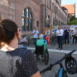 Teilnehmer einer mobilen Bürgerversammlung im Juli 2022 stehen im Kreis um die Redner OB König und Planungs- und Baureferent Daniel F. Ulrich. Im Vordergrund steht eine Teilnehmerin mit dem Rücken zur Kamera neben ihrem Fahrrad. Vor ihr sitzt eine Frau im Rollstuhl.