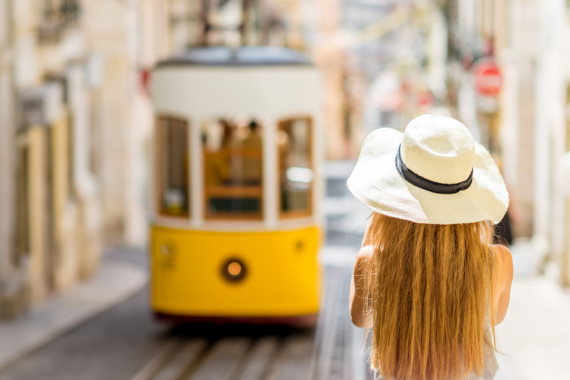 Frau mit blonden langen Haaren mit Strohhut und Sommerkleid die eine alte gelbe Straßenbahn im Sonnenschein fotografiert.