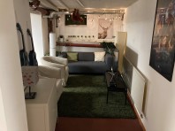 Im Kellerraum steht ein weißes Sofa auf einem dunkelgrünen Teppich. An der Decke sind Heizungsrohre, an der Wand hängen zwei kleine blaue Ukulelen.