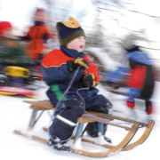 Ein Kind in einem Schneeanzug, das auf einem Holzschlitten sitzt und einen Hügel herunterfährt.