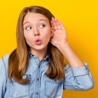 Ein Teenager-Mädchen hält die Hand hinter ihr linkes Ohr, blickt nach links und lauscht aufmerksam. Sie trägt ein Jeanshemd und steht vor einem neutralen, gelben Hintergrund.