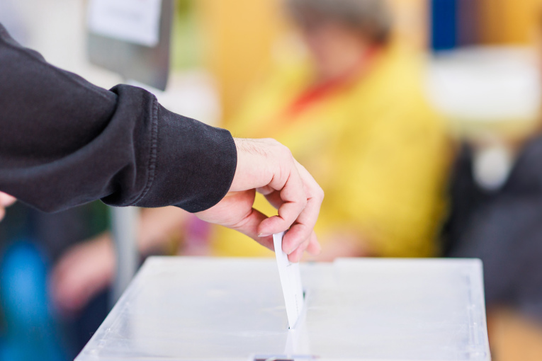 Eine Hand hält einen Wahlzettel und steckt diesen in den Schlitz einer Wahlurne.