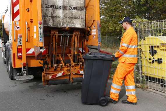 Foto von einem Müll·mann mit einer Rest·müll·tonne. Der Müll·mann schiebt die Müll·tonne zu einem Müll·auto.