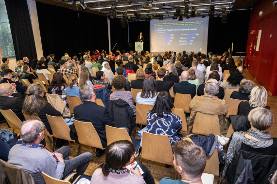 Foto: Viele Menschen sitzen in einem Tagungs·raum und hören einem Vortrag zu.
