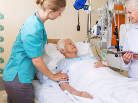 Das Bild zeigt ein Zimmer in einem Kranken·haus. In dem Bett liegt eine alte Frau. Neben dem Bett stehen 2 Frauen vom Kranken·haus.