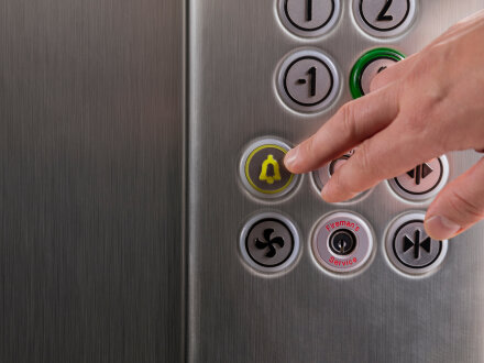 Das Bild zeigt eine Hand und die Tasten in einem Aufzug. Die Hand ist über der Taste für: Not·ruf.
