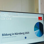 Auf zwei Bildschirmen sind Statistiken zu Bildung zu sehen, von hinten ist eine Person mit halblangen Haaren zu sehen, sie sitzt vor den Bildschirmen.