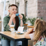 Mother and little daughter eating in the street cafe. Trendy hipster Mother with dreadlocks and toddler daughter having lunch together at cafe summer day.
