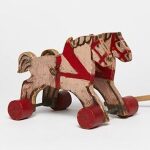 Man sieht alte Spielzeugpferde aus Holz neben einer hölzernen Spielzeugkutsche.