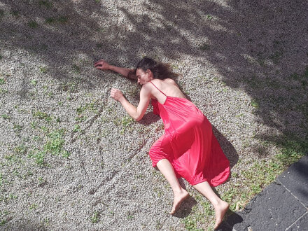 Foto von einer Frau in einem roten Kleid. Die Frau liegt auf dem Boden. Der Boden ist aus Kies., Bild © Susanna Curtis
