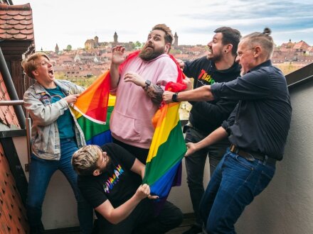 Foto von 5 Personen auf einem Balkon. Die Person in der Mitte hat eine Regen·bogen·fahne um die Schultern gelegt. Die anderen 4 Personen ziehen an der Regen·bogen·fahne., Bild © Keckos