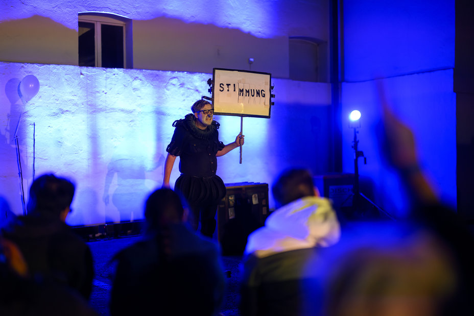 Ein dunkel gekleideter Clown auf einer blau beleuchteten Bühne hält ein schwarz umrandetes Schild mit der Aufschrift "Stimmung" hoch., Bild © (c) Steffen Kirschner l viaframe / Steffen Kirschner | viaframe