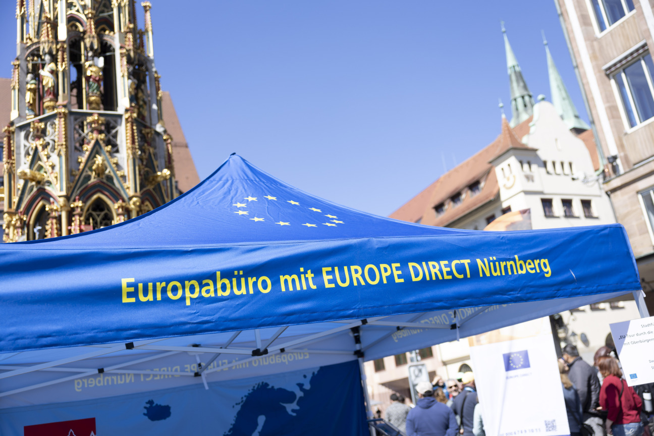 Das Bild zeigt ein blaues Zelt auf dem in gelber Schrift Europabüro mit EUROPE DIRECT Nürnberg geschrieben steht. Im Hintergrund ist der schöne Brunnen zu sehen.