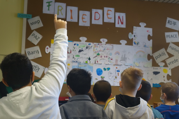 Die Schulkinder stehen vor verschiedenen Motiven für ihr Friedenspuzzle und wählen per Meldung eines aus