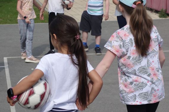 Zwei Mädchen halten sich an den Händen, eines der Mädchen hat in der anderen Hand einen Fußball.