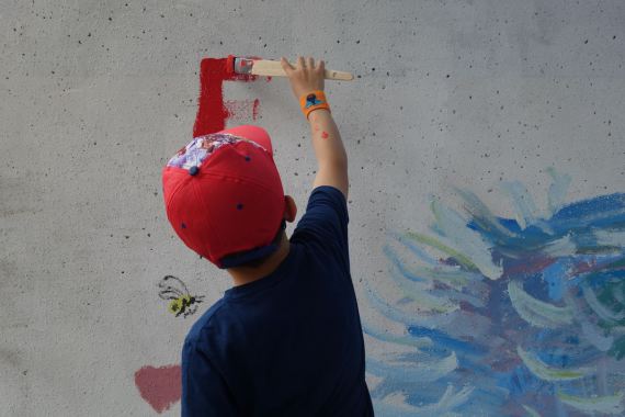 Ein Junge mit roter Käppi malt einen Buchstaben mit einem Pinsel an eine Wand.