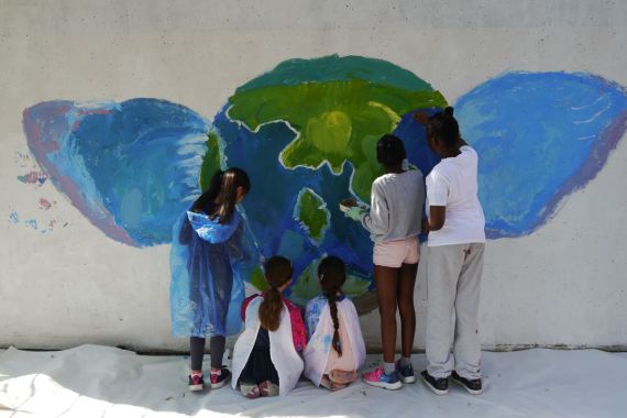 Kinder malen gemeinsam eine Weltkugel mit Flügeln auf eine Wand im Pausenhof.