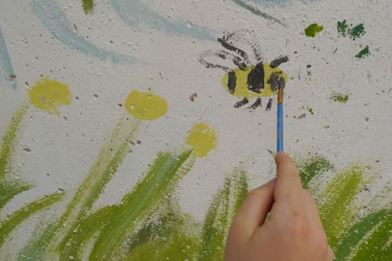 Eine Hand hält einen feinen Pinsel und malt eine Biene auf eine Wand.