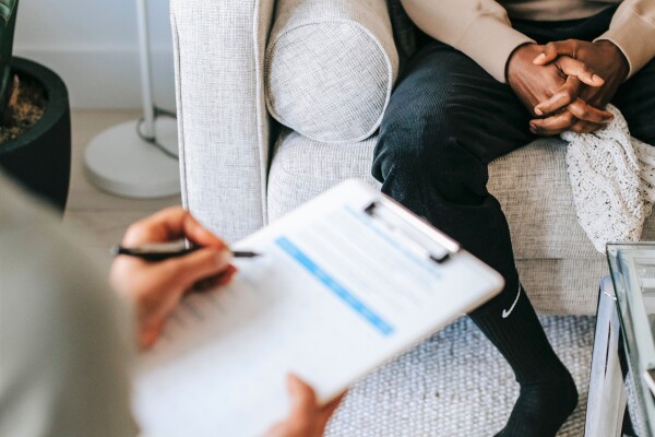 Eine Frau mit Klemmbrett sitzt auf einem Stuhl. Ihr gegenüber sitzt jemand auf einer Couch mit verschränkten Händen.