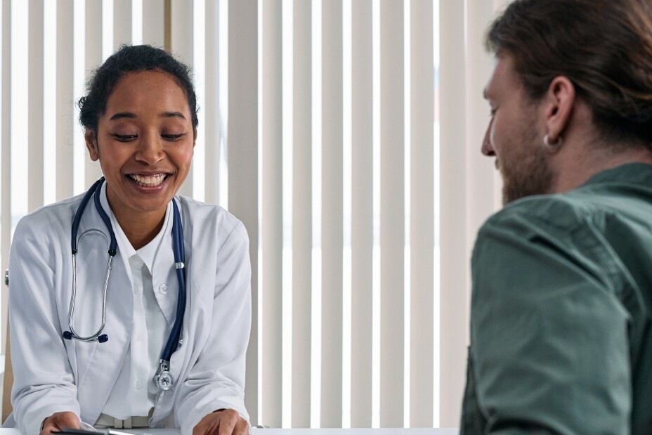 Eine Ärztin mit einem Klemmbrett in der Hand bespricht sich mit einem Mann ihr gegenüber an einem Tisch.