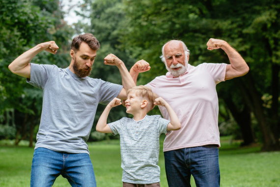 Männer aus drei Generationen stehen nebeneinander auf einer Wiese und zeigen spielerisch ihre Muskeln.