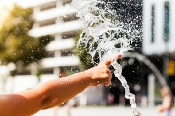 Ein Wasserstrahl spritzt auf einem gepflasterten Platz vor einem Hochhaus in die Luft, ein nackter Arm steckt den Finger in den Wasserstrahl.