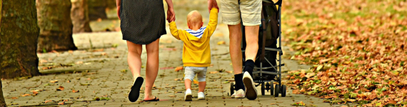Mutter und Vater im Park halten Kleinkind zwischen sich an den Händen.