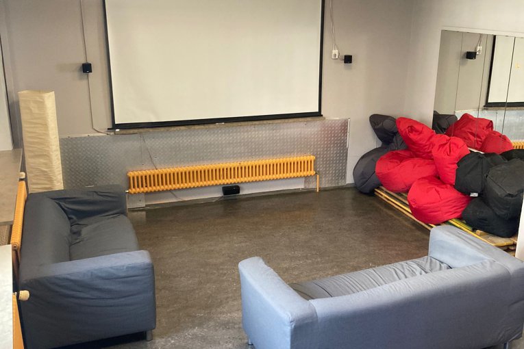 Zwei graue Sofas stehen vor einer weißen Leinwand. An der Decke hängt ein Videobeamer. Im Hintergrund sind rote und schwarze Sitzsäcke aufgestapelt.