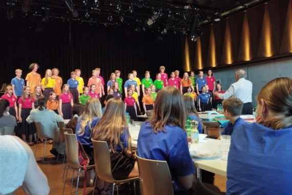 Kinder singen. Sie stehen in bunten T-Shirts auf einer Bühne im Großen Saal. Kinder in blauen T-Shirts sitzen an Tischen und schauen zur Bühne.