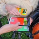Drei Personen scannen im Kreis auf einer Wiese stehend mit ihrem Smartphone einen QR Code vom jeweils anderen Smartphone ab, um an einer Musik Installation teilzunehmen.