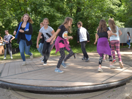 Foto von einer Balance∙scheibe. Eine Gruppe von Kindern läuft und balanciert auf der Scheibe. Eine Mitarbeiterin vom Erfahrungs·feld ist auch dabei., Bild © Stadt Nürnberg/Christine Dierenbach