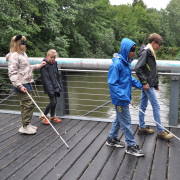 Vier Kinder mit Langstöcken und Augenbinden laufen über die Brücke.