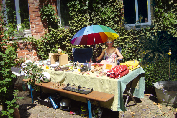 Eine Frau im Sommerkleid und mit Sonnenbrille sitzt an einem Flohmarkttisch in einem Gartenstuhl. In der einen Hand hält sie einen Regenbogen-Regenschirm. Auf dem Tisch und der dazugehörigen Bierbank liegen verschiedene Gegenstände.