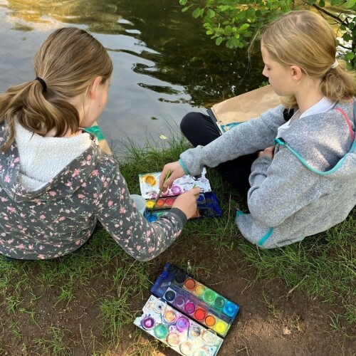 Zwei Kinder sitzen in der Natur, neben einem Fluss, und malen mit Wasserfarben die Landschaft.