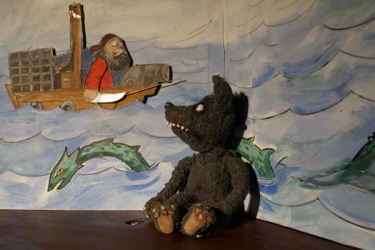Eine Wolfpuppe sitzt auf der Bühne. Im Hintergrund ein Bild vom Meer in dem Gefahren lauern, eine Seeschlange und ein Boot