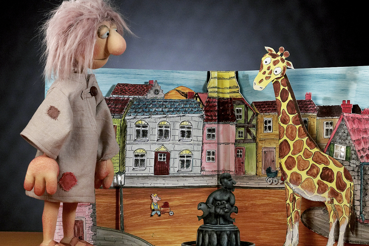 Zwei puppen vor dem Bild eines Dorfes. Links ein Riese und rechts eine Giraffe zwischen ihnen ein Brunnen, Bild © Dornerei Puppentheater