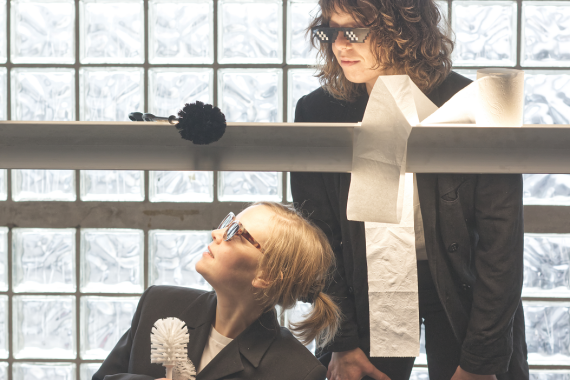 Zwei Menschen im Agenten Outfit mit Sonnen Brille und vor ihnen eine rolle Klopapier und eine Klobürste