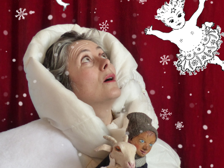 Foto von einer Frau in einer dicken weißen Jacke. Die Frau steht vor einem roten Vorhang. An sich gepresst hält die Frau 2 Figuren: ein Kind und eine Ziege. Die Frau schaut nach oben., Bild © Amrey Haage / Figurentheater Weidringer