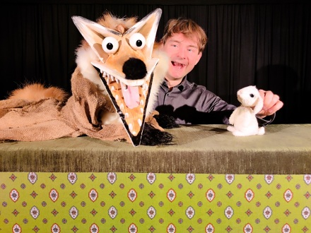 Foto von einer Hand·puppe. Die Hand·puppe sieht aus wie ein Fuchs. Ein Mann hält die Hand·puppe. Daneben sitzt ein kleiner weißer Hase. Der Hase schaut den Fuchs an., Bild © Theater DERenDINGEn@Uli Voland
