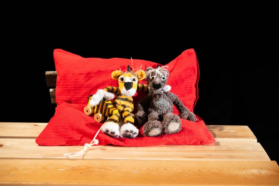 Tiger und Bär sitzen Arm in Arm auf einem großen roten Kissen. T