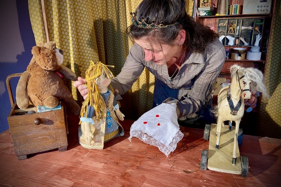 Eine Frau steht in einer altmodischen Puppenwerkstatt. Ein Teddy in einer Holzkiste, ein Holzpferd und eine Puppen sind neben der Frau zu sehen. Mit der Puppen scheint sie zu sprechen.