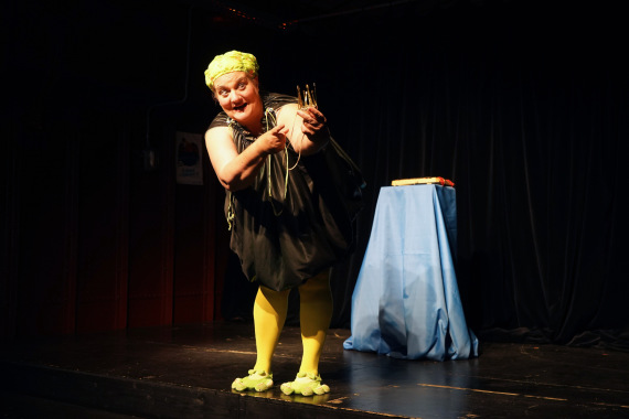 Auf einer dunklen Bühne steht eine als Frosch verkleidete Frau und deutet mit dem Finger auf eine Krone, die sie in der anderen Hand hält. Im Hintergrund steht ein mit blauem Stoff verhülltes Podest.