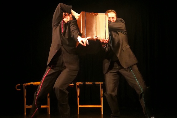 Zwei Personen in dunklen Anzügen rangeln mittels zweier Koffer miteinander.
