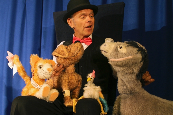 Ein Mann in schwarzem Anzug, Hut und roter Fliege, sitzt vor einem dunkelblauen Vorhang. Auf dem Schoß hat er drei Kuscheltier: Katze, Hund und Hahn. Neben ihm steht ein Esel-Kuscheltier, mit dem er im Gespräch zu sein scheint.