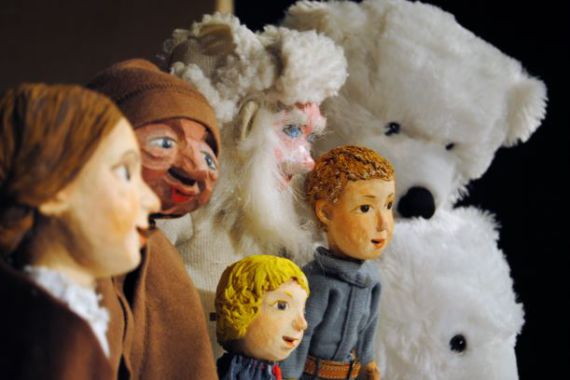 Vor einem dunklen Hintergrund sind mehrere Handpuppen zu sehen. Im Vordergrund zwei Erwachsenen in brauner Kleidung, im Bildmittelpunkt ein Mann mit weißem Bart und weißer Fellmütze und zwei Kinder in blauen Pullovern. Dahinter zwei Eisbären.