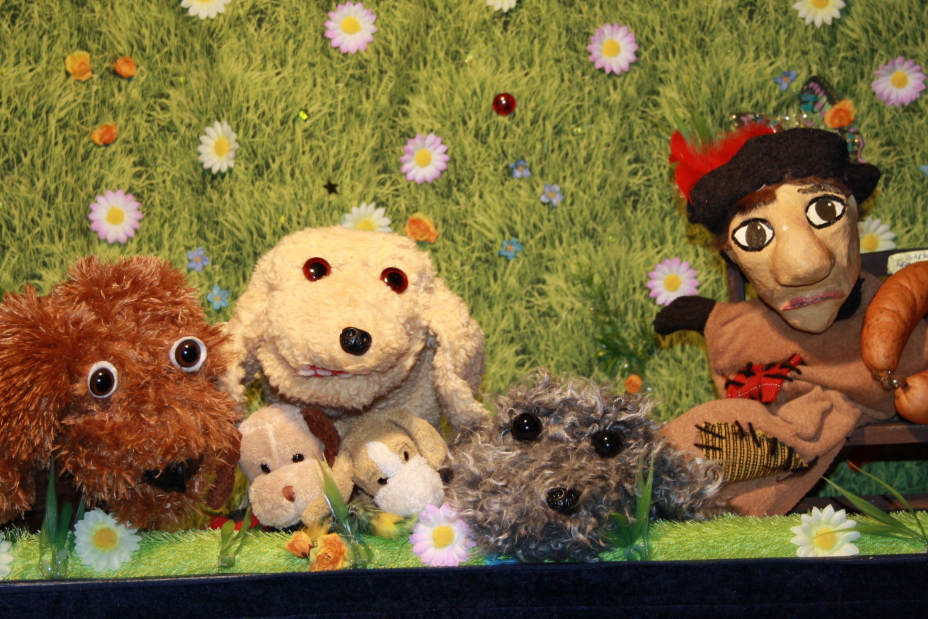 Fünf Hunde kuscheln zusammen neben einer Bank, auf der der Kasper mit einer großen Wurst sitzt., Bild © Krowis Puppenbühne