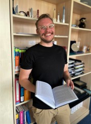 Matthäus Wilga steht vor einem Regal und hält ein aufgeschlagenes Buch in der Hand