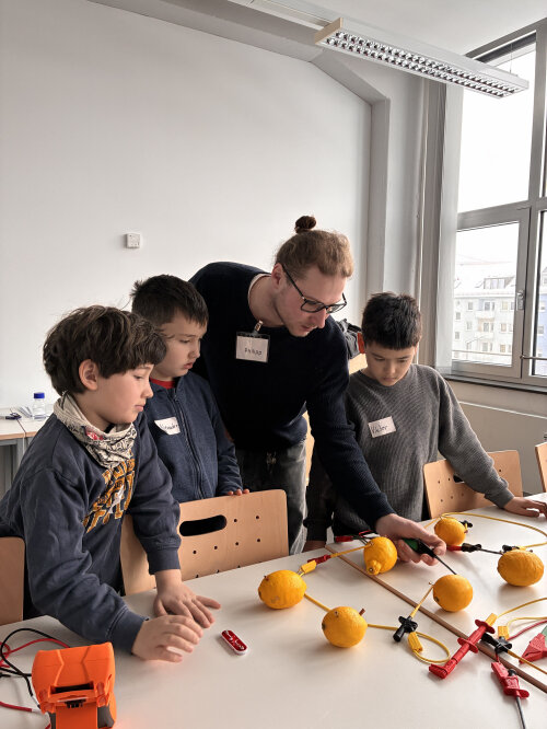 Drei Kinder schauen einem Erwachsenen bei einem Experiment zu, bei dem aus einer Zitrone Strom gewonnen wird.