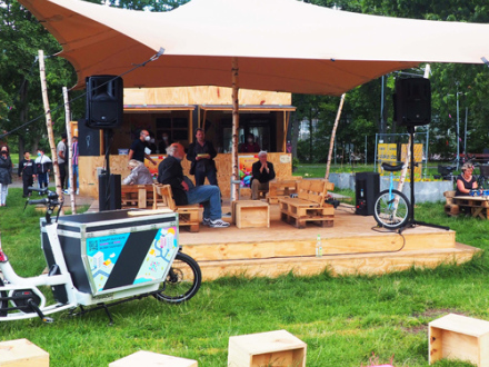 Foto vom mobilen Kultur·laden KommVorZone im Anna·park. Auf einer Wiese steht ein Zelt. Unter dem Zelt steht eine Bühne aus Holz. vor der Bühne stehen Holz·würfel zum Sitzen., Bild © Olga Komarova / Stadt Nürnberg